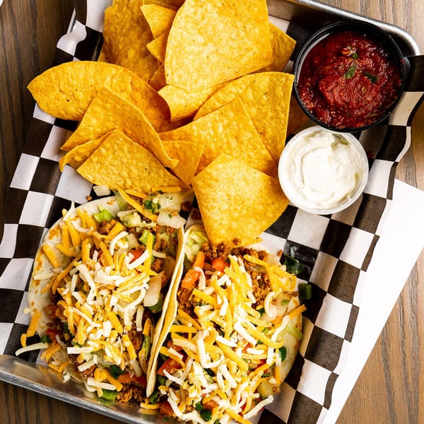 Two Rookies Sports Bar & Grill tacos with chips, salsa, and sour cream on a metal tray.
