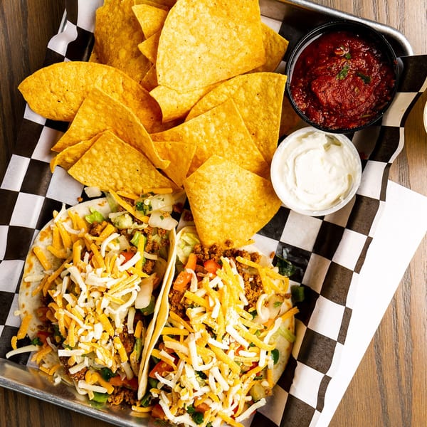 Two Rookies Sports Bar & Grill tacos with chips, salsa, and sour cream on a metal tray.