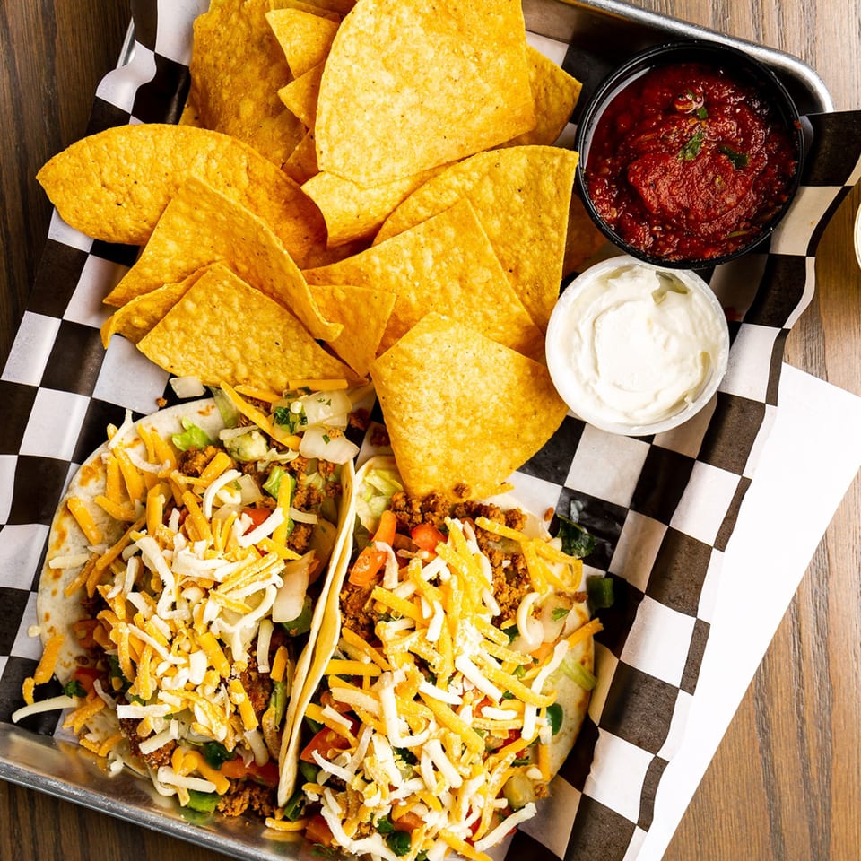 Two Rookies Sports Bar & Grill tacos with chips, salsa, and sour cream on a metal tray.