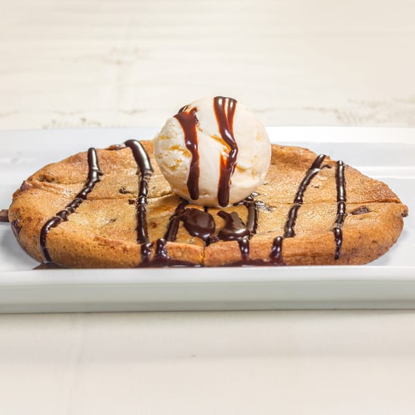 Warm chocolate chip cookie topped with vanilla ice cream and chocolate syrup on a white plate.