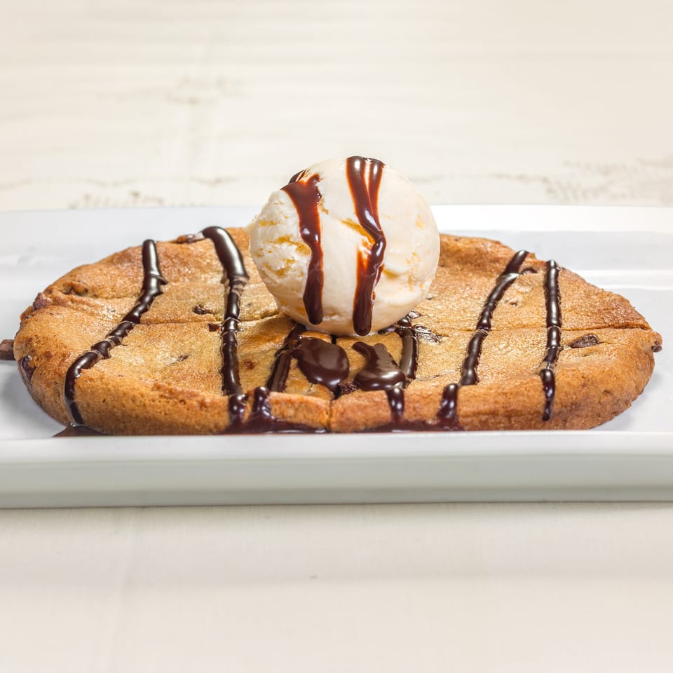 Warm chocolate chip cookie topped with vanilla ice cream and chocolate syrup on a white plate.
